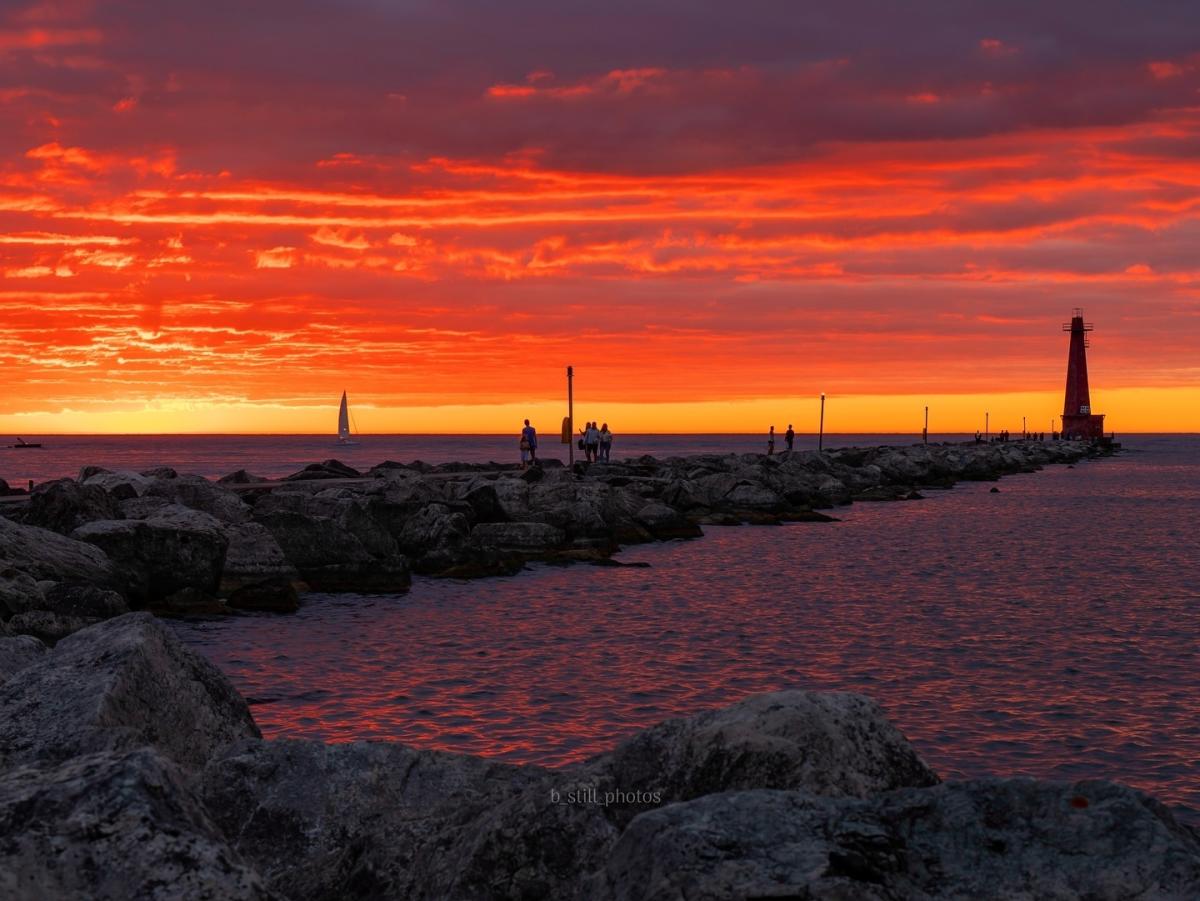 Sunset at a Pere Marquette Park in Muskegon featuring a fiery sky and clouds over the calm waters of Lake Michigan, with a lighthouse and several people on a rocky breakwater.