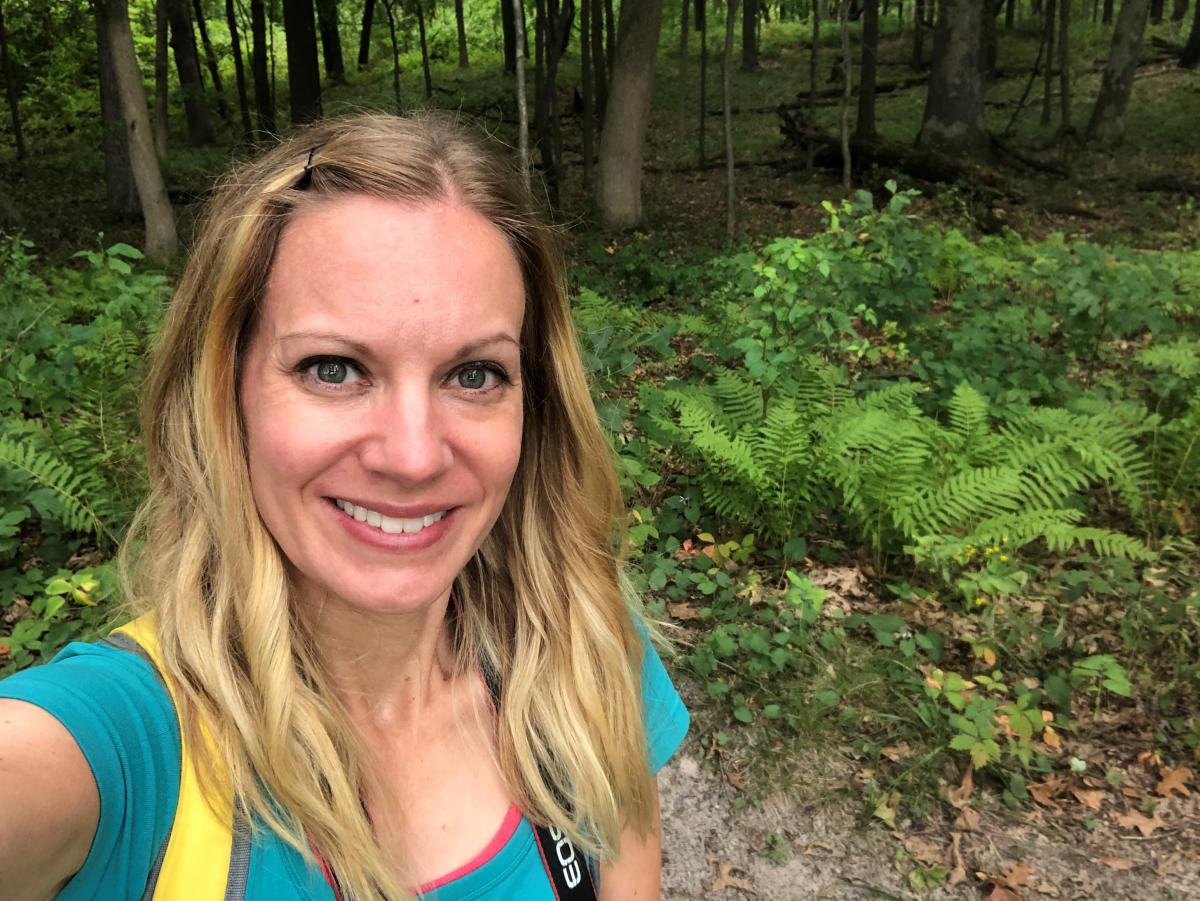 A blonde woman poses in front of vegetation in the woods. She is wearing an aqua shirt.