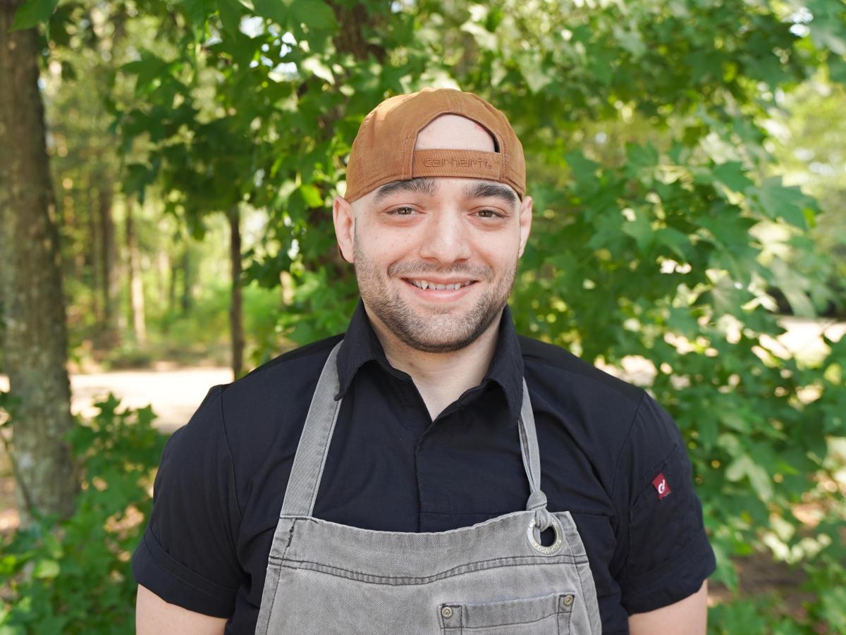 Chef Bryce Grenier of Palmettos on the Bayou smiles outdoors in his chef coat, gray apron and cap, with leafy green trees in the background.
