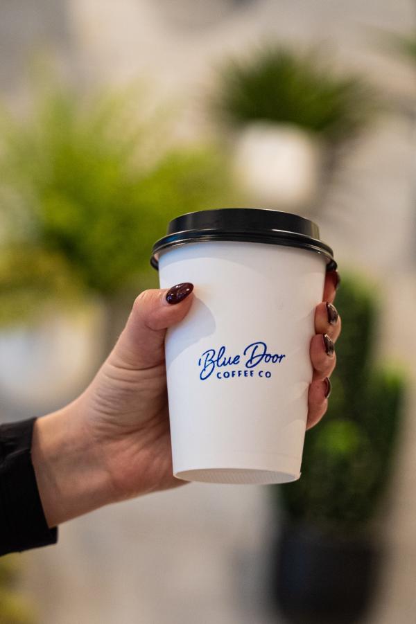 A woman with burgundy nails holds a white paper cup that reads, "Blue Door COFFEE Co." (in blue text) in front of a wall lined with potted plants.