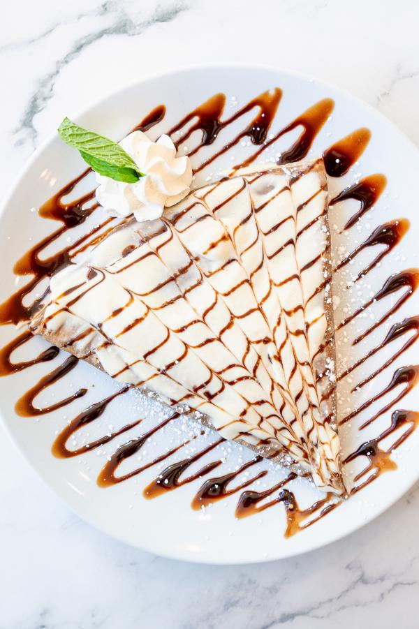 A close-up shot of a tiramisu crepe coated in cream cheese, drizzled in chocolate syrup, and garnished with whipped cream and a mint leaf.