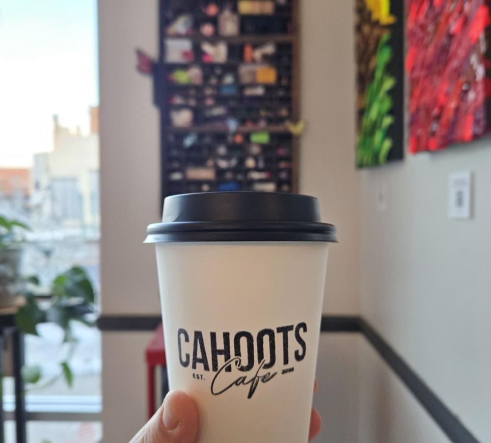 Person holds a coffee to-go cup that reads "Cahoots Cafe". In the background is a blurry view of a treasure board, filled with small trinkets, and two paitings hang on a nearby wall.