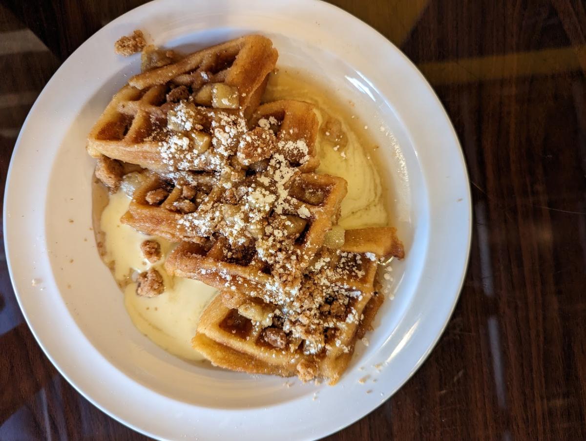 Image is of 4 golden waffles with apples, cinnamon crunch bits, powder sugar and a cream sauce.