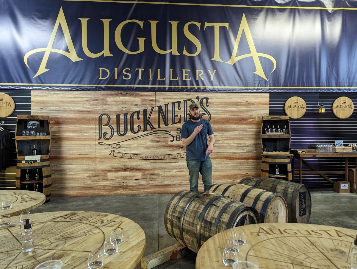 A bearded man speaking surrounded by bourbon barrels and a sign that says Augusta Distillery