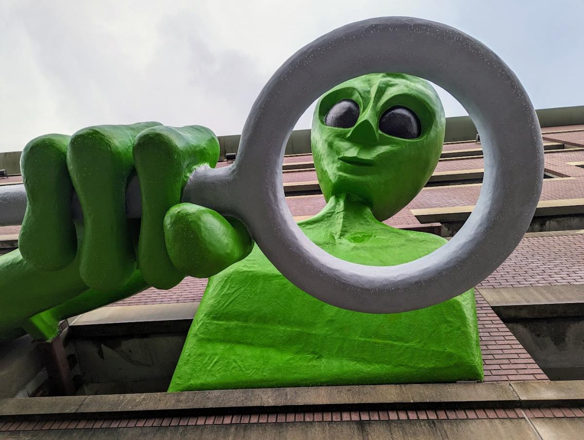 A 30 foot green alien leaning out of a parking garage peering down through a giant magnifying glass