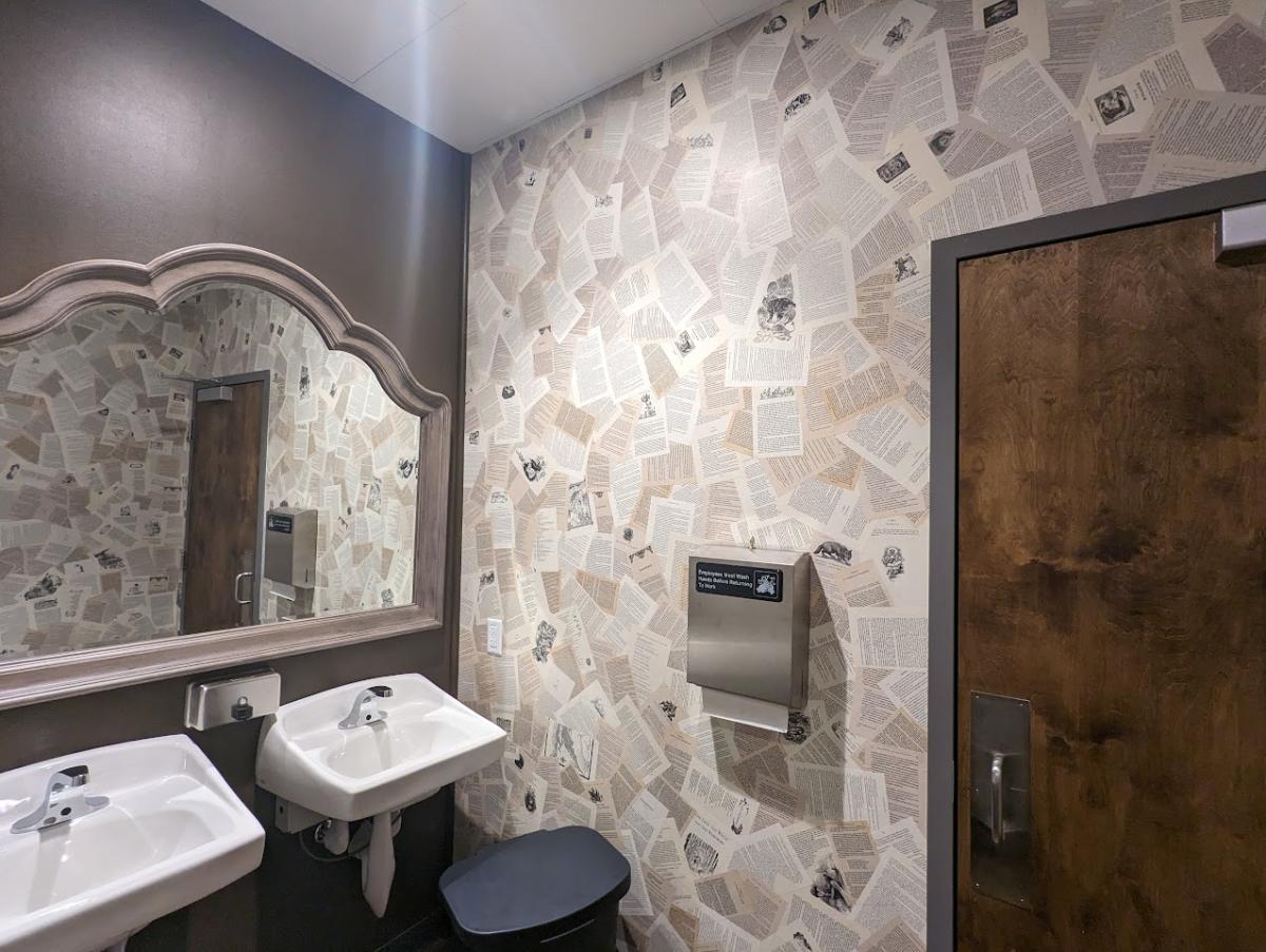 Inside the bathroom where you can see a mirror, two sinks, towel dispenser and wall paper that looks like pages from a book.