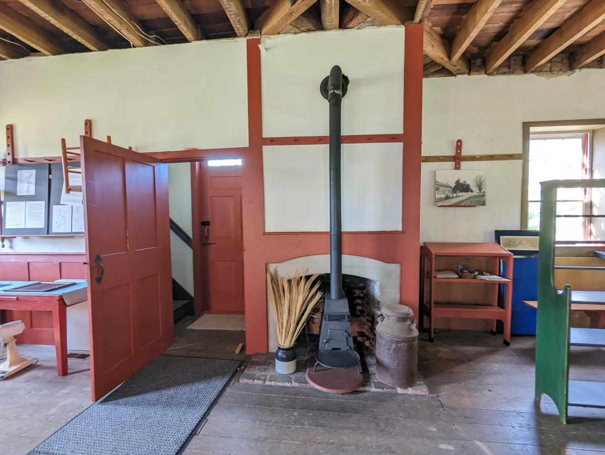 Image is of inside the Meeting House with a stove against the wall and a peg rail that runs along the room on the wall.
