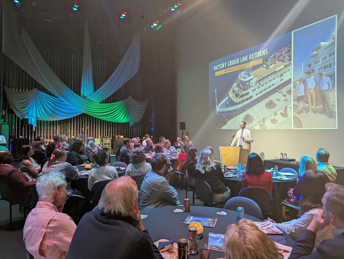 a room is full of seated people attentively listening to a speaker in front of a large projection screen