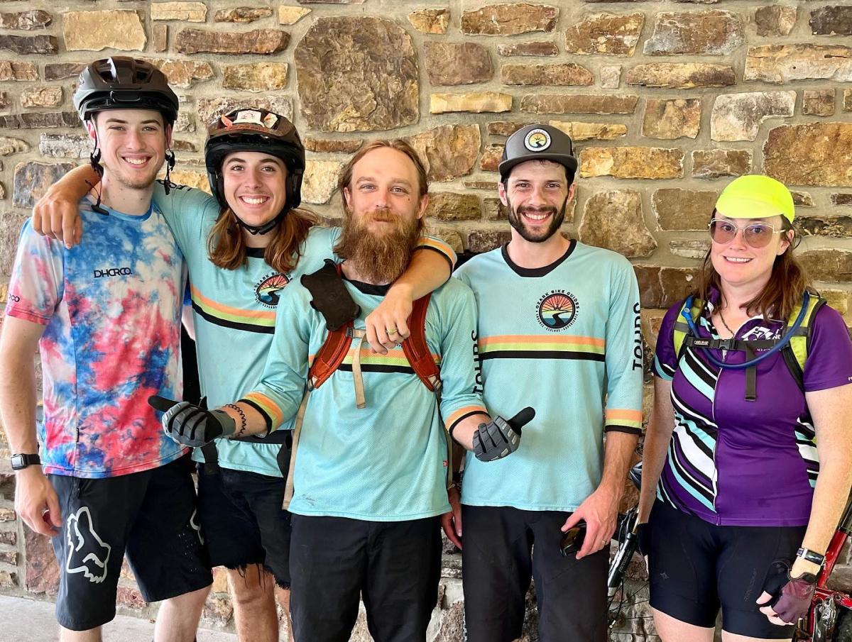 Five smiling individuals, likely mountain bikers, pose in front of a textured stone wall, wearing cycling attire and appearing in good spirits.