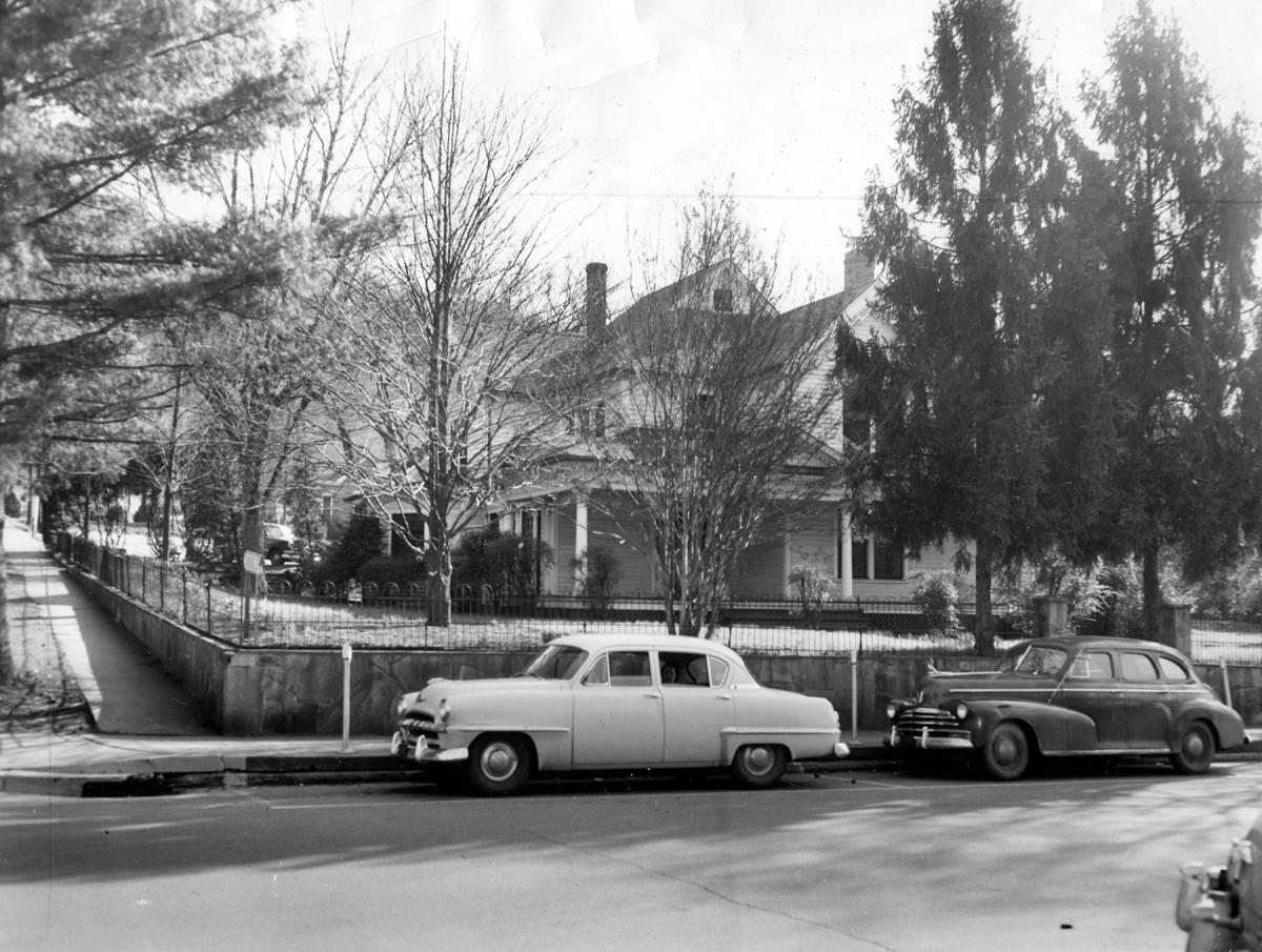 The McKee family home behind an iron fence and trees at Main and Landis streets in Sylva, NC, around 1952, with two 1950s cars parked in front