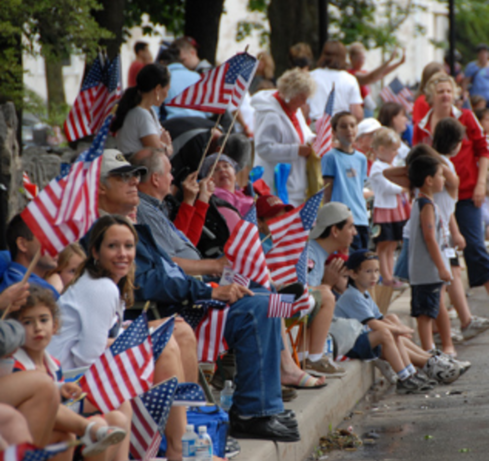 A diverse group of onlookers watch the Skippack Fourth of July Parade from the sidewalk. Numerous spectators are waving American Flags and wearing variations of red, white, and blue.