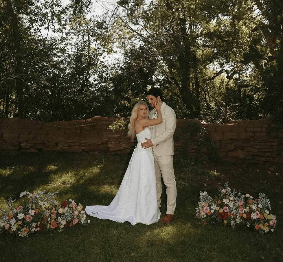 A bride in a white gown and a groom in a beige suit embrace in a sunlit garden, surrounded by colorful floral arrangements.