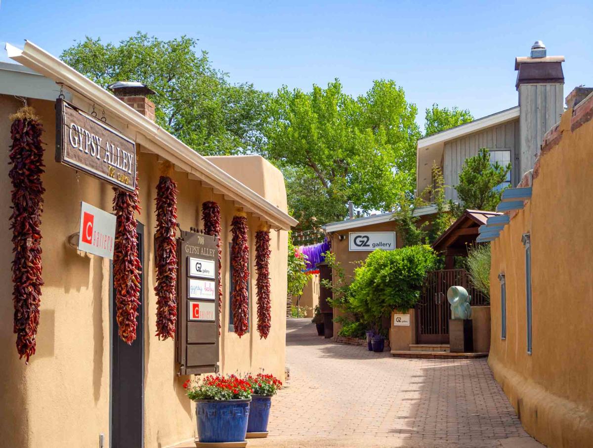 View of a sunny street in Santa Fe, featuring traditional adobe architecture adorned with hanging chili ristras. Signs indicate locations such as "Gypsy Alley" and "G2 Gallery." Flowerpots add colorful accents along the pathway.