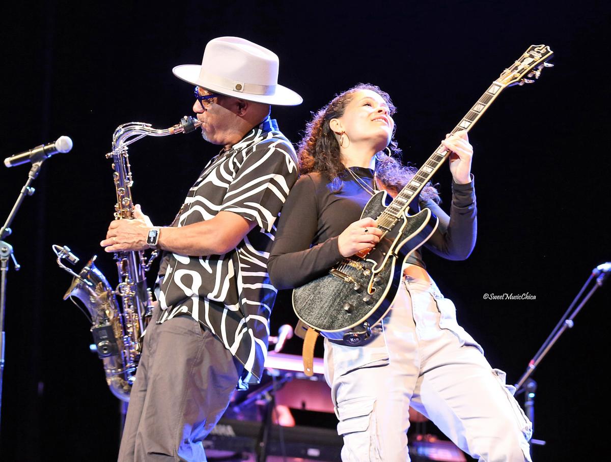 Jazz musicians Kirk Whalum and Andréa Lisa perform together on stage. Kirk Whalum, wearing a white wide-brimmed hat and patterned black-and-white shirt, plays the saxophone with energy. Andréa Lisa, wearing a black top and light cargo pants, leans back with a joyful expression while playing an electric guitar. The stage is dimly lit with spotlights, highlighting the passion and connection between the two performers.