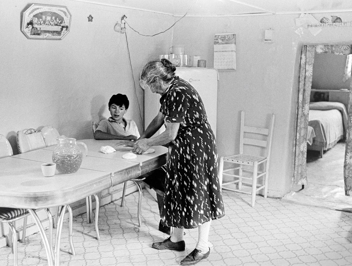 Black and white photograph of a woman preparing tortillas at a kitchen table while a child watches nearby.