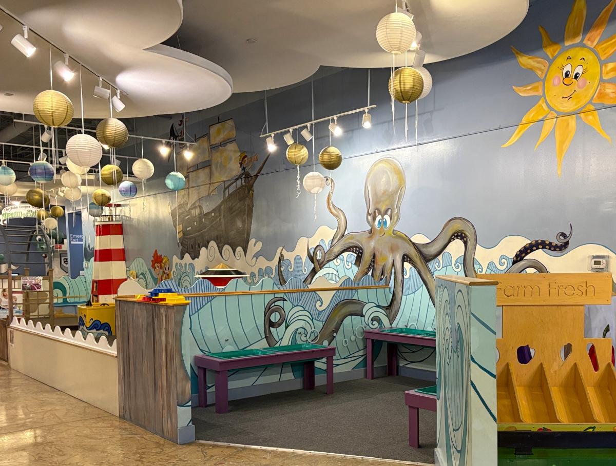 The camera focuses on a play area within The Woodlands Children's Museum. The walls are painted with cartoon-like waves, a pirate ship, a giant octopus, and a smiling sun. In the back, there is a red-and-white striped lighthouse. On the right, there is part of a farmer's market exhibit visible.