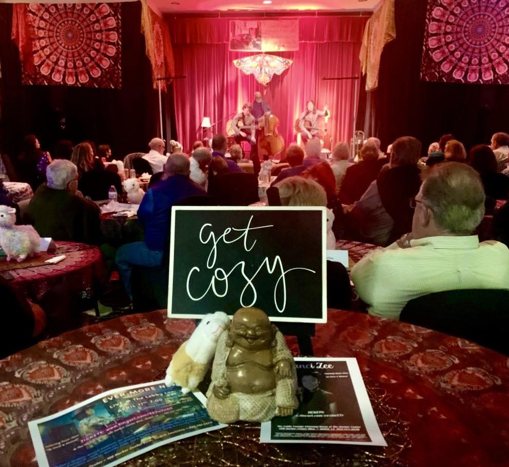 Audience seated at tables during a Lobby Lounge concert, watching musicians perform on a warmly lit stage beneath red curtains, with a “Get Cozy” sign in the foreground.