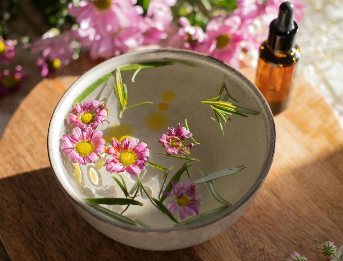 Pink flowers and fresh herbs floating in a ceramic bowl beside an amber dropper bottle.