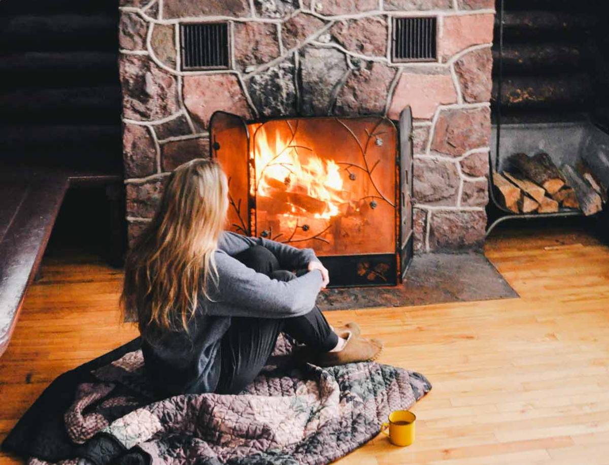 Girl sits beside stone fireplace with warm drink.