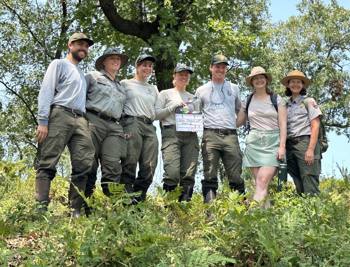 A group of seven people, including park rangers and a visitor, pose together on a grassy hillside surrounded by lush greenery and trees. Most of them wear khaki and green uniforms with wide-brimmed hats, while one person in the group wears a light-colored skirt and a backpack. One individual in the center holds a clapperboard, suggesting a film or documentary project. They are all smiling under a bright blue sky, standing in a natural outdoor setting.