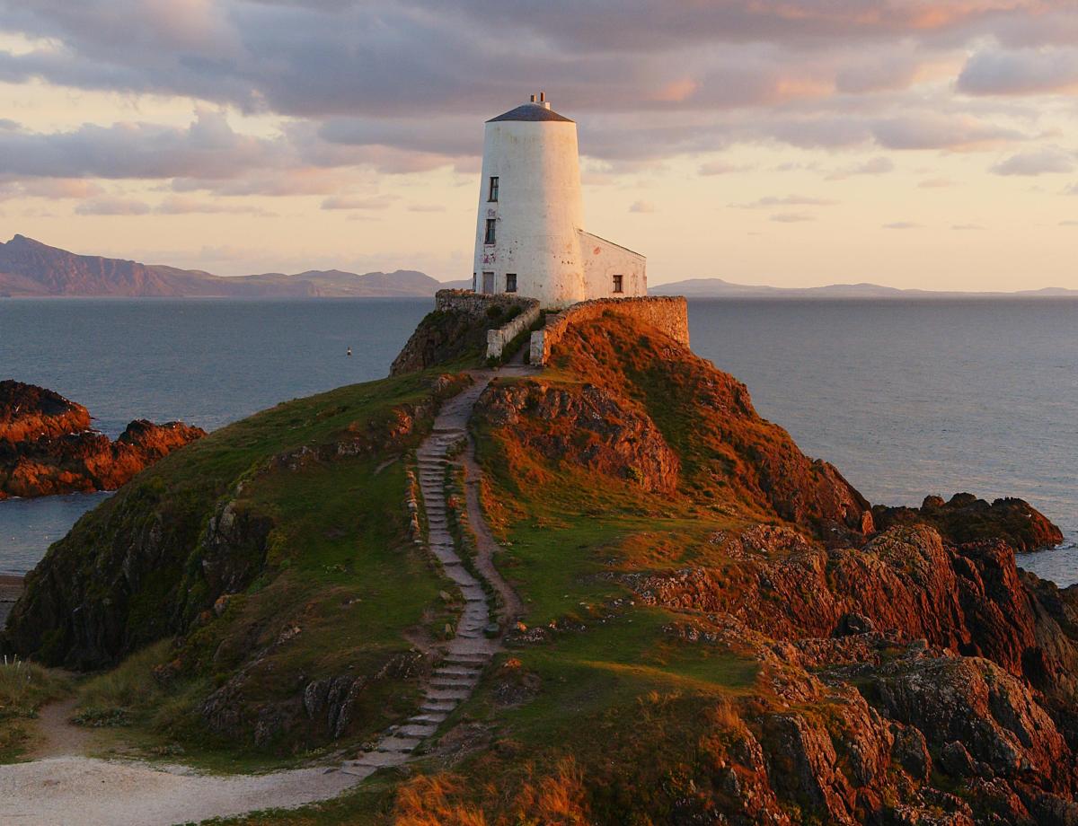 White lighthouse-style building on a rocky headland, reached by a narrow stone path, overlooking the sea at sunset, with warm golden light on the cliffs and distant mountains visible across the water beneath a partly cloudy sky.
