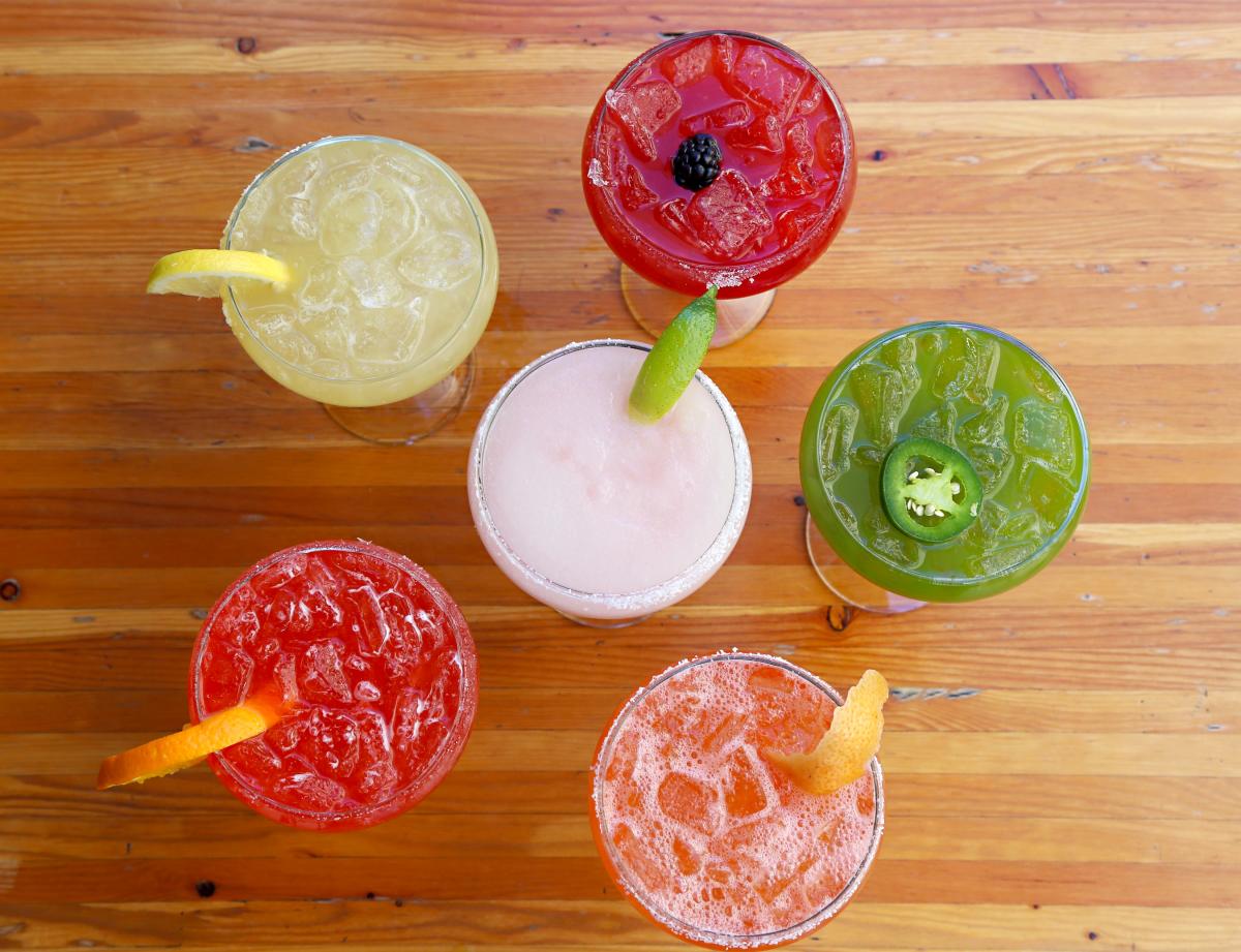5 margaritas all different colors with different garnishes surrounding another margarita glass. The view is from above and the glasses are sitting on a wood table.