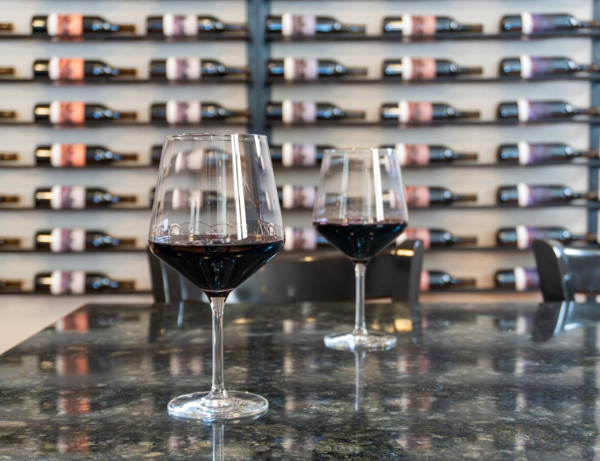 Two glasses of red wine in wine glasses sit on a countertop. A wall of colorfully-labeled wine bottles lay horizontally on a wall rack in the background, slightly out of focus.