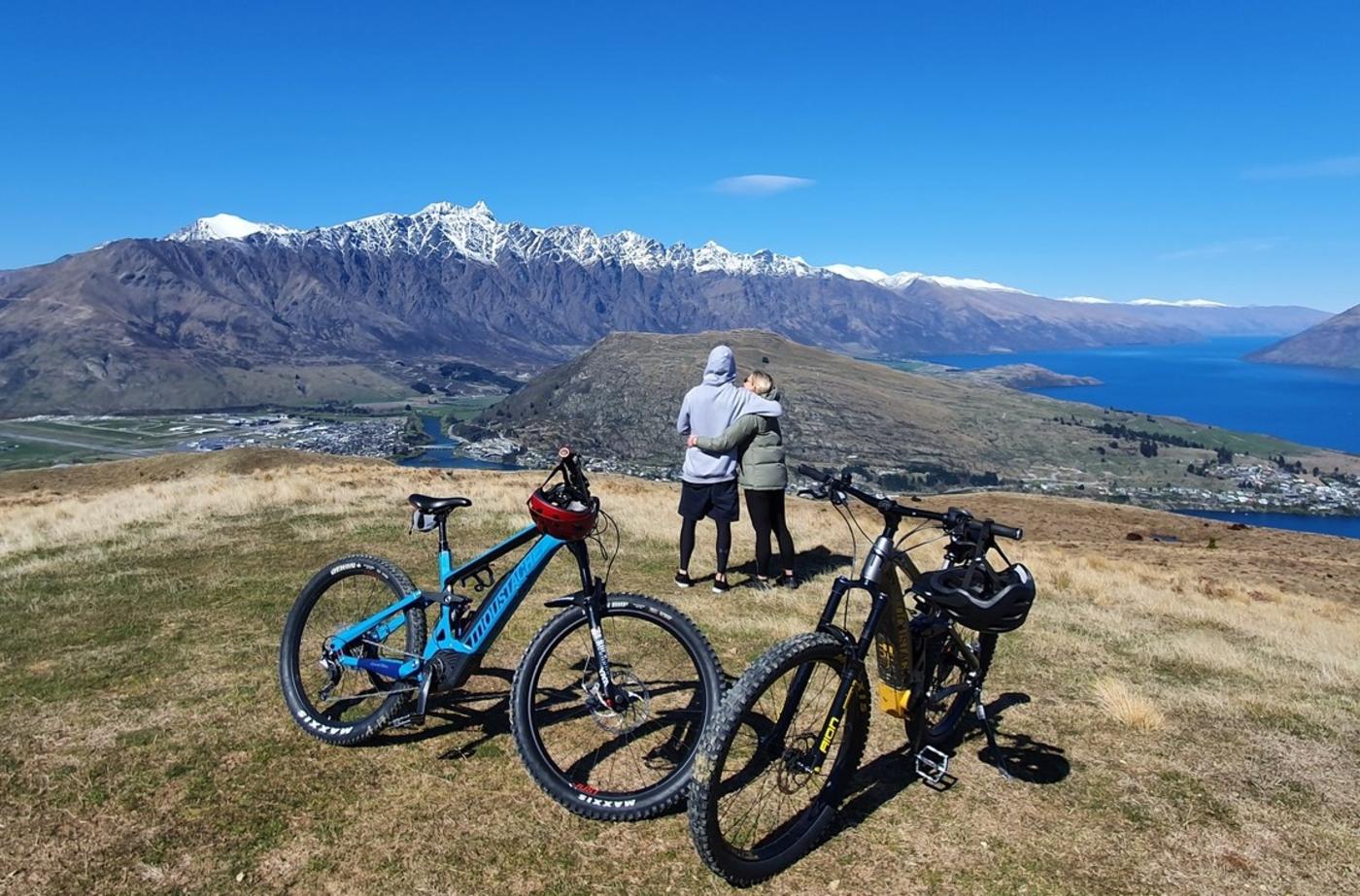 Two people with bikes standing on a mountain overlooking snowy peaks and lake