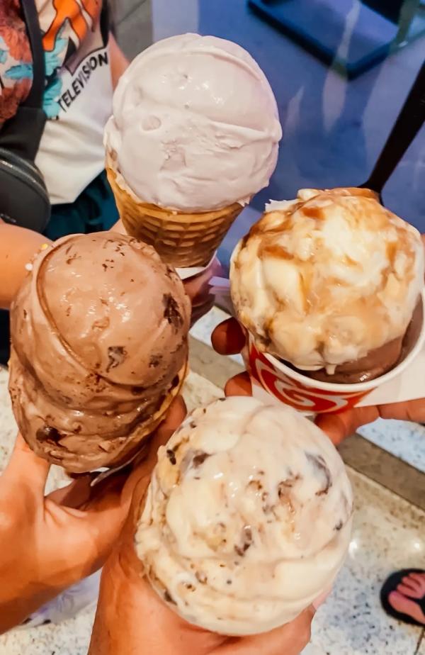 Image of four scoops of ice cream. Three scoops are in waffle cones and one is in a cup. Four separate people are holding the scoops in the center of the picture.