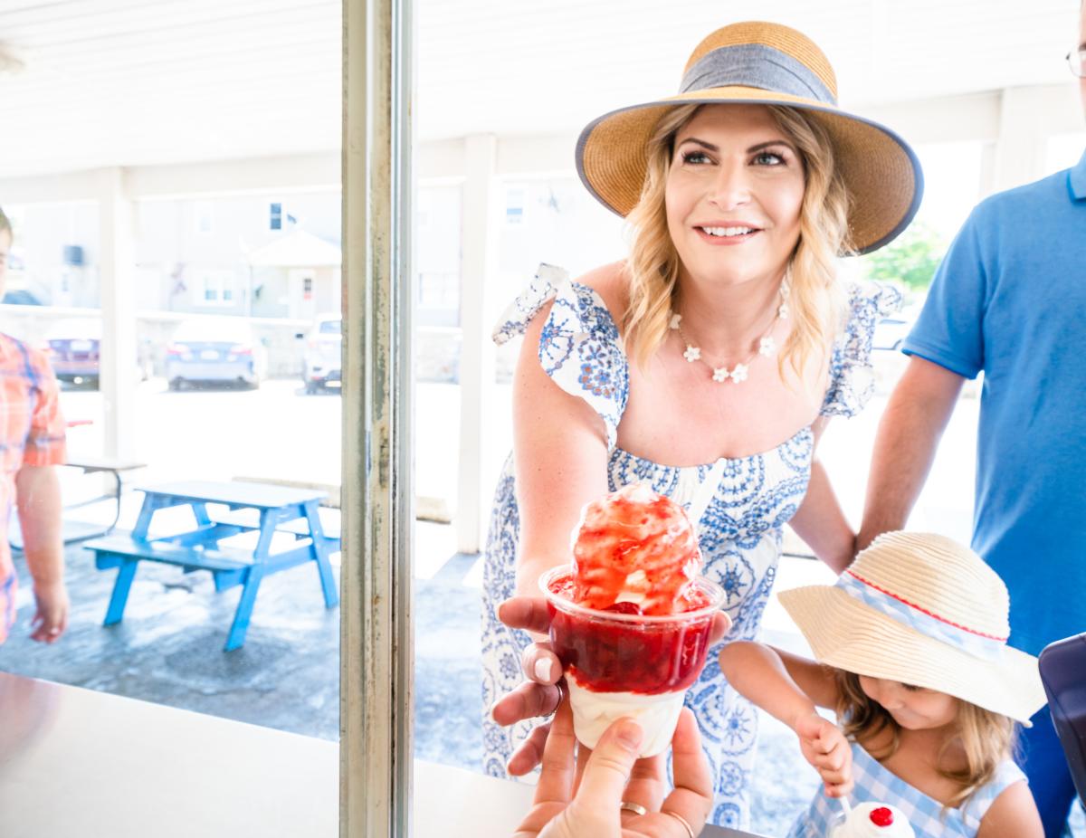 A woman in a large brim hat is handed a strawberry sundae out the window of an ice cream shop.