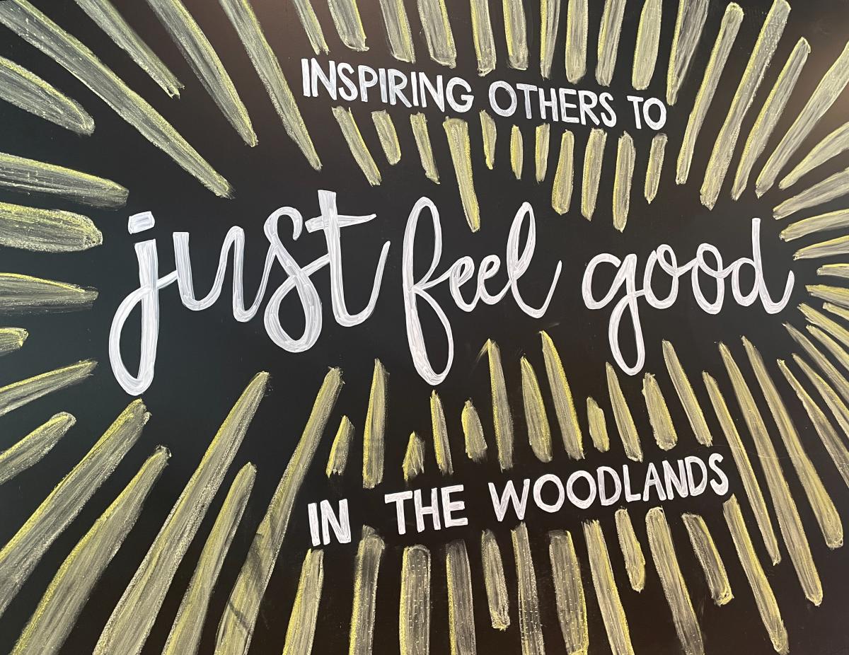 A black chalkboard reads "INSPIRING OTHERS TO just feel good IN THE WOODLANDS" in white text. The text is surrounded by lots of bright yellow stripes to look like a sun or a flower.
