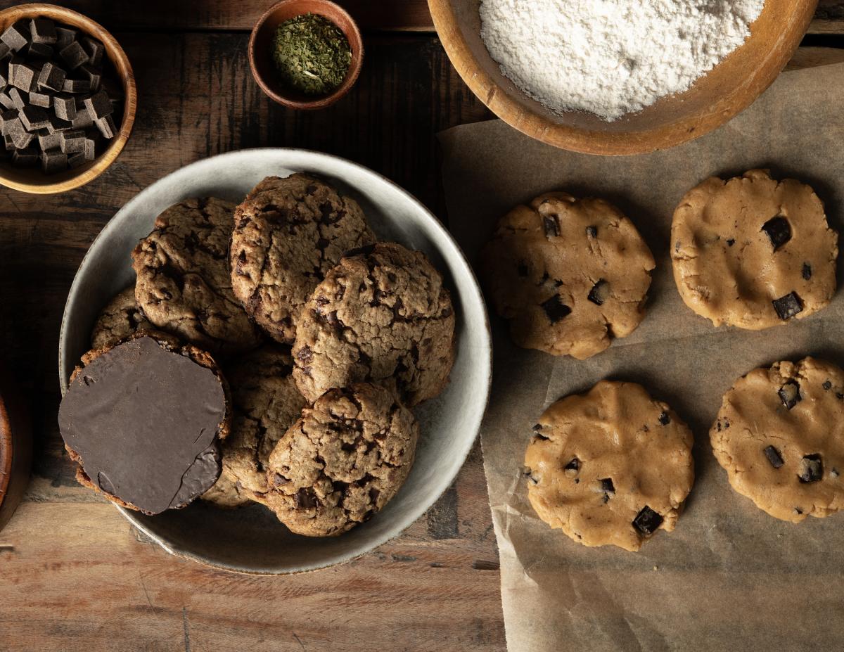 A bowl of chocolate chip cookies next to raw cookie dough on parchment paper. Nearby are bowls of flour, chocolate chunks, and herbs on a wooden table.
