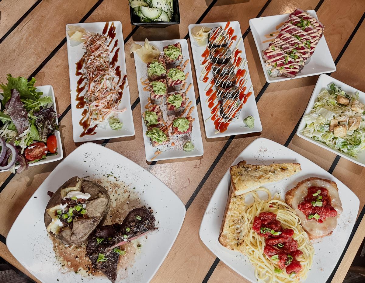 A table full of food plates, including sushi, steak, spaghetti, and more.