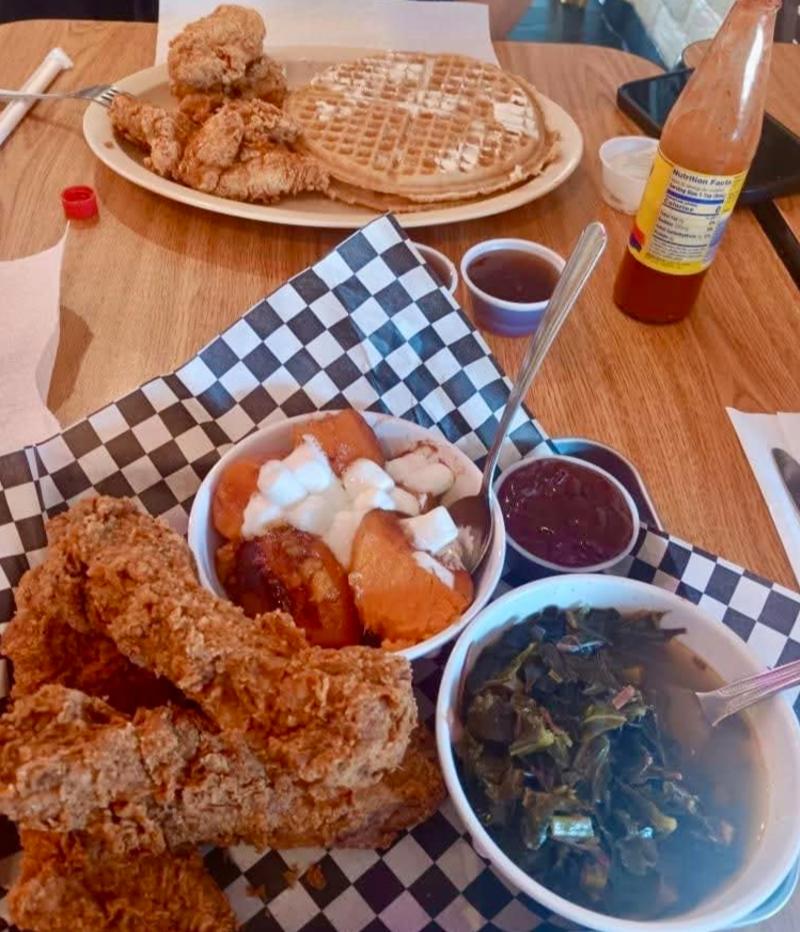 A photo of a table at Frank's Famous Chicken and Waffles. The closest plate features fried chicken, sweet potatoes and greens. The plate farther away is a chicken and waffles plate.