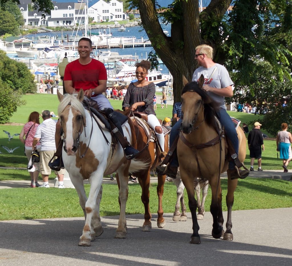 A group of three people ride horseback up a hill on Mackinac Island with the marina in the background