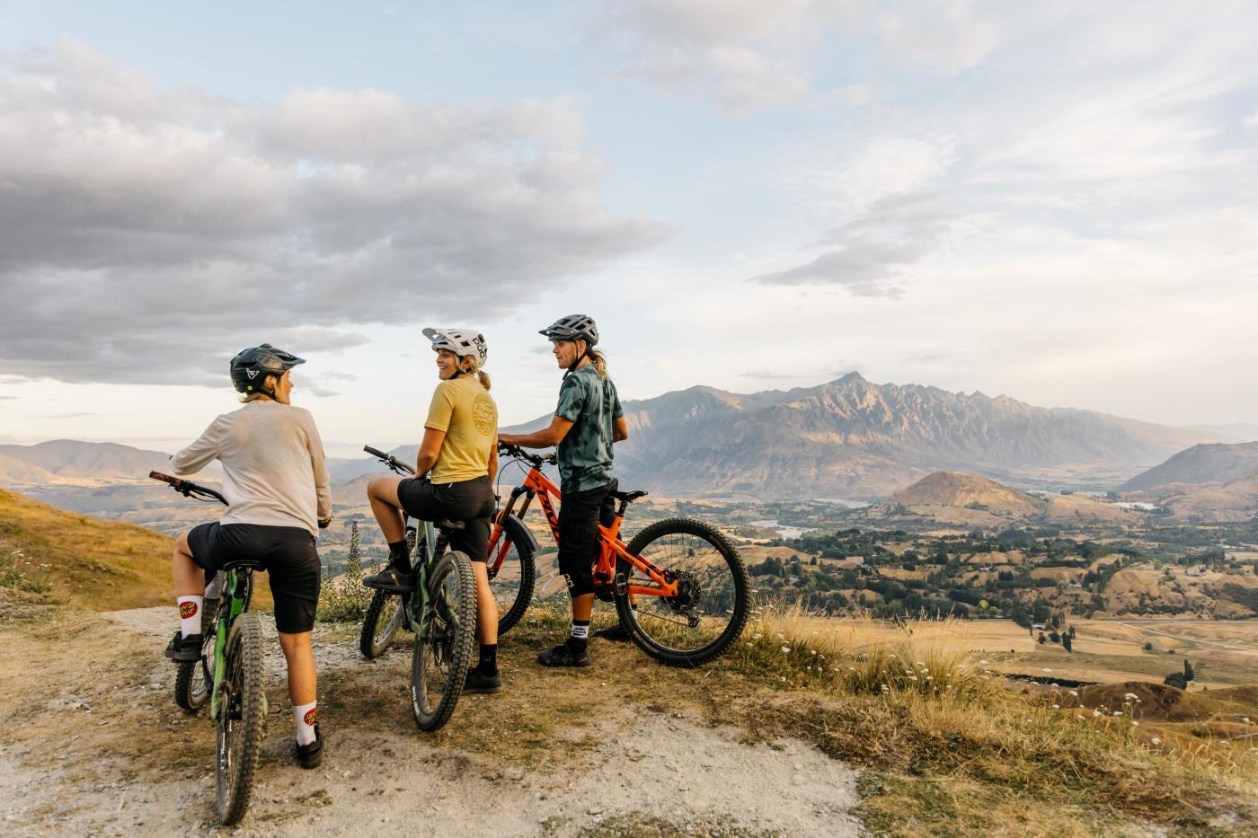Three mountain bikers at the top of a mountain overlooking a valley with the Remarkables mountains in the background