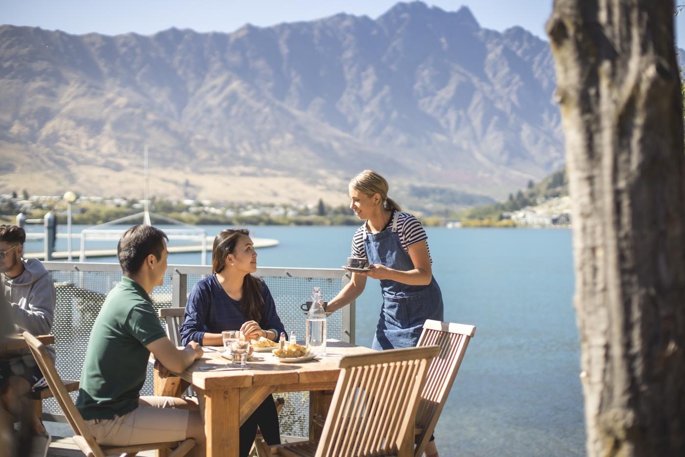 Couple being served by a waitress outside at the Boat Shed café, with the Remarkables Mountains in the background