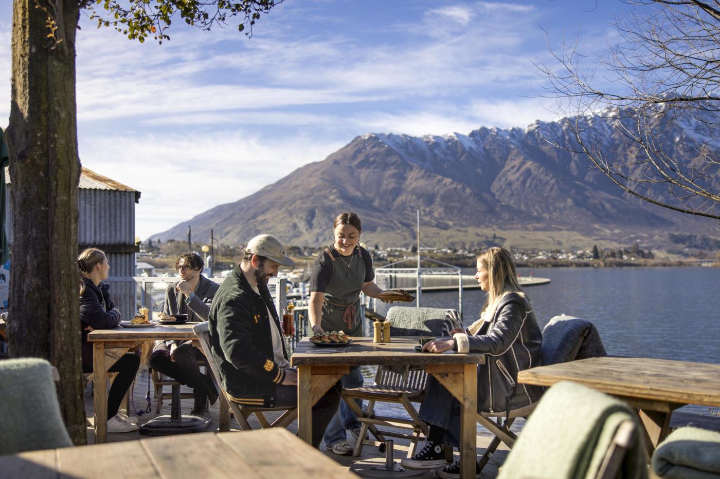 People dining outside next to the lake with the Remarkables mountain range in the background at The Boat Shed