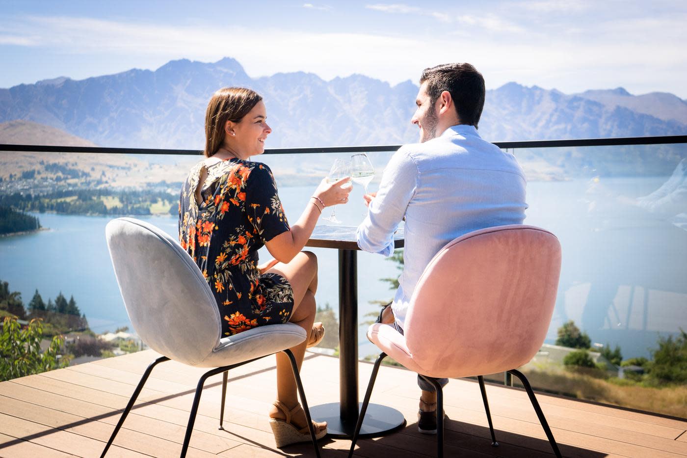 A couple enjoying a drink on The Nest Restaurant and Bar outdoor deck overlook the lake and the Remarkables Mountains