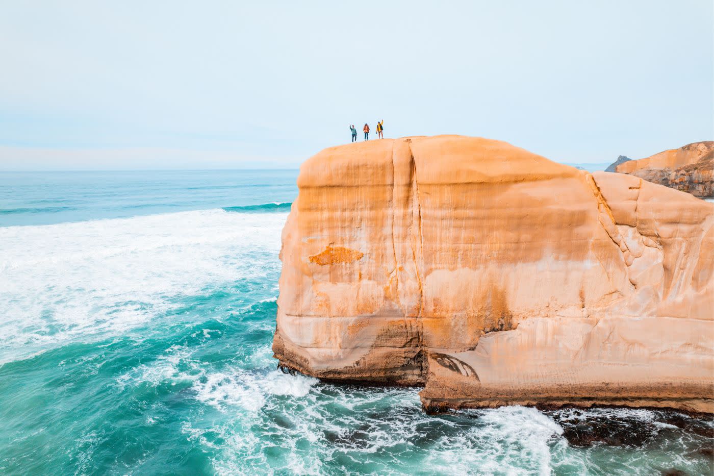 Three people stand on cliff overlooking ocean at Tunnel Beach near Dunedin