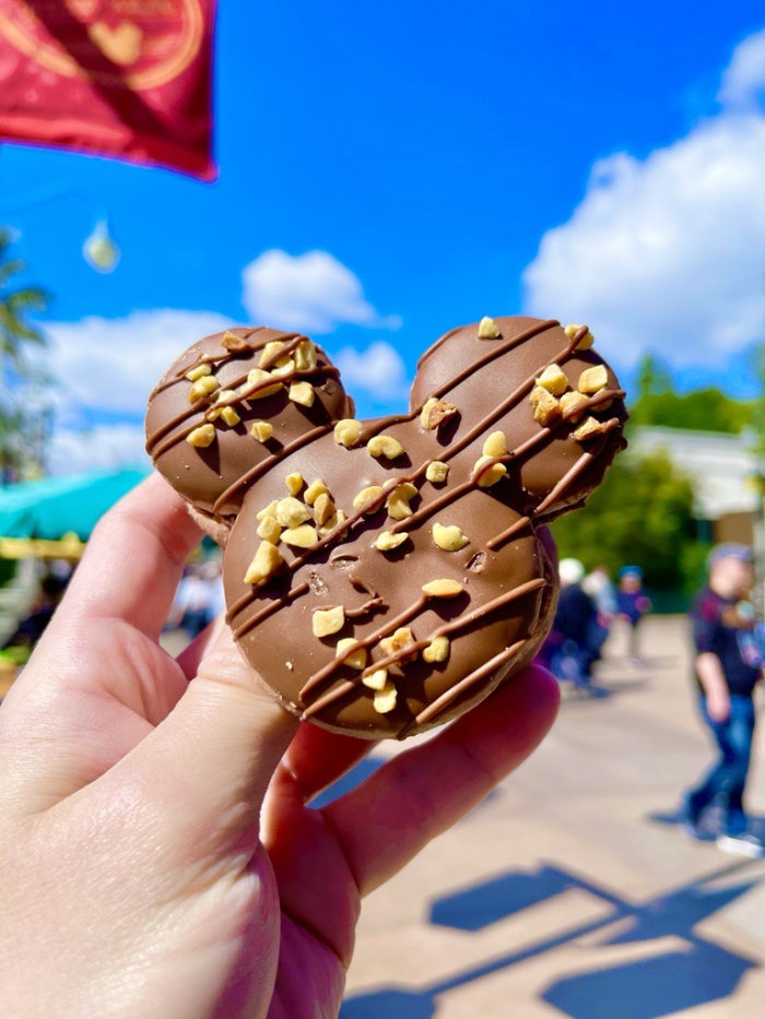 Image of a Mickey Mouse-shaped macaron being held up against the sky. The cookie is chocolate covered with a chocolate drizzle and nuts.