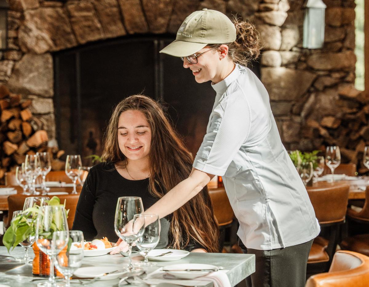 A smiling server in a white uniform and green cap places a plate of food in front of a happy customer seated at a set dining table. The warm, rustic background features a massive stone fireplace filled with stacked wood.