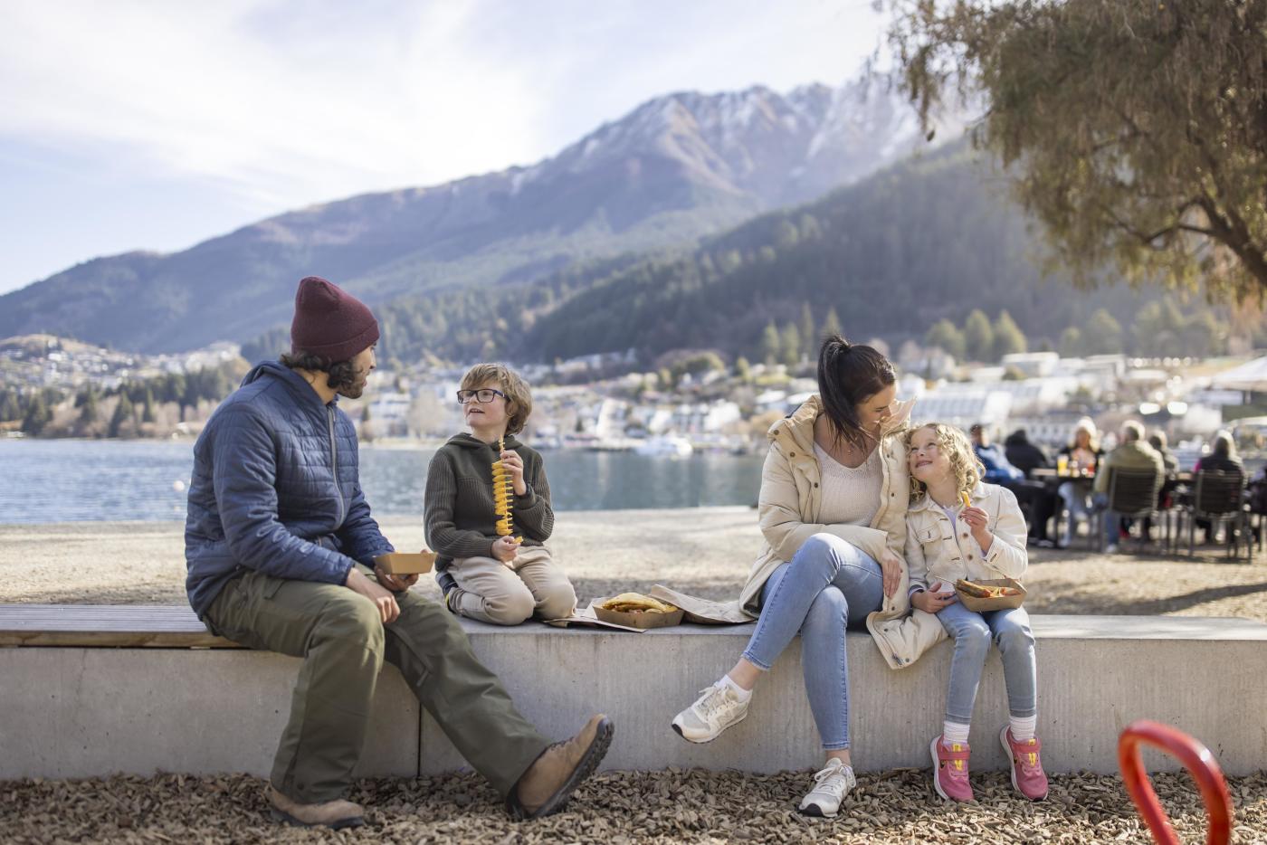 Family enjoying fish and chips by Queenstown waterfront with mountains in the background
