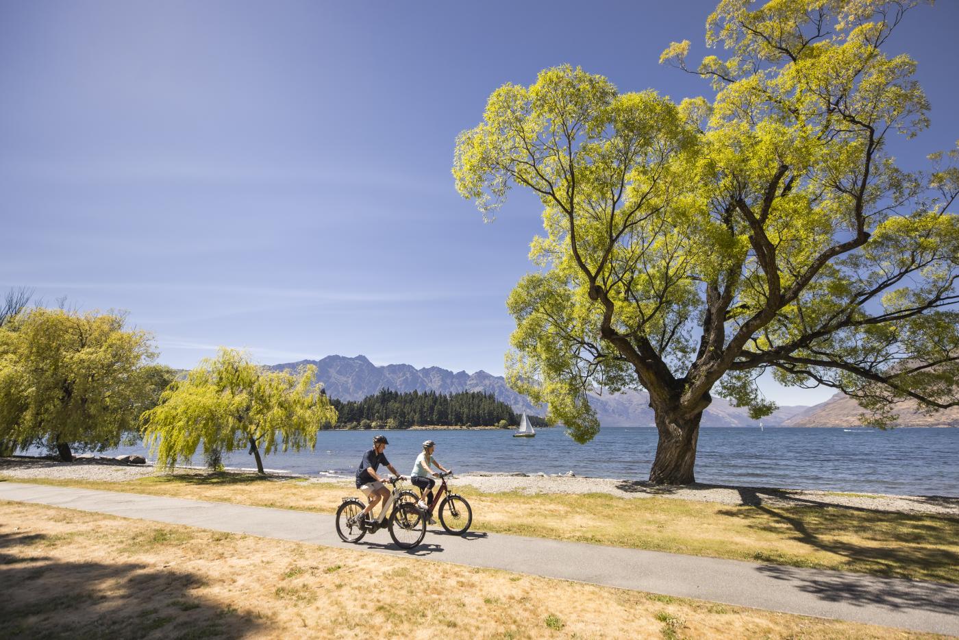 A couple bike along Queenstown Waterfront with the Remarkables mountain range in the background on a sunny day