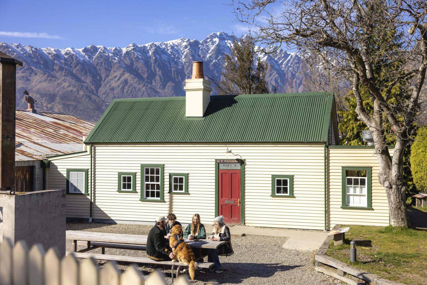 People eating food in an outside courtyard infront of a cute cottage cafe with mountains in the background