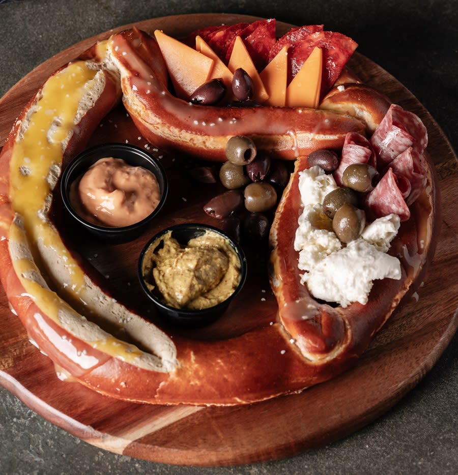 A large, round, soft pretzel on a wooden platter with two dipping sauces, assorted pepperonis, and olives