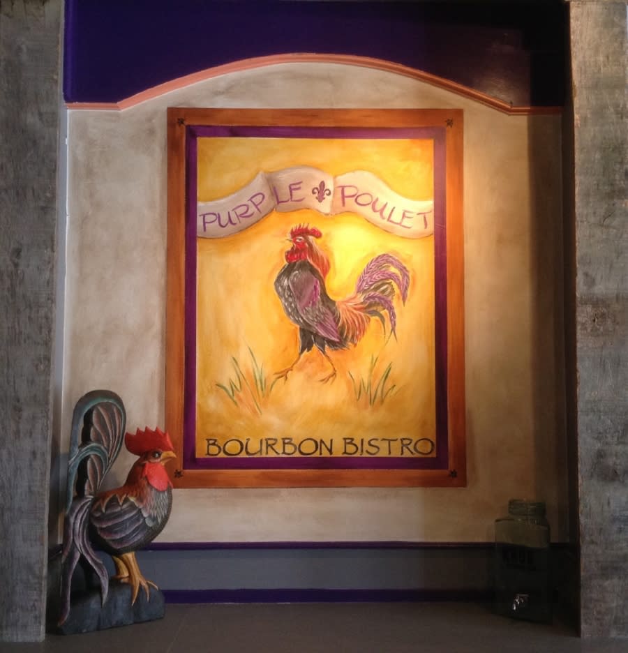 A wooden rooster in the lower corner and a painting with the words purple poulet bourbon bistro painted around a purple rooster