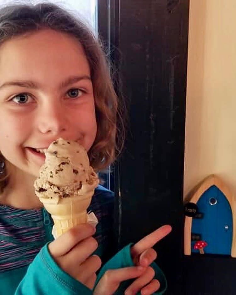 Little girl holding ice cream cone in front of her smile while pointing at a blue fairy door on the fairy door trail.
