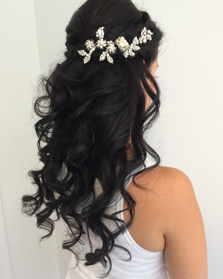 A woman stands with long black curls and a crown of flowers and pearls