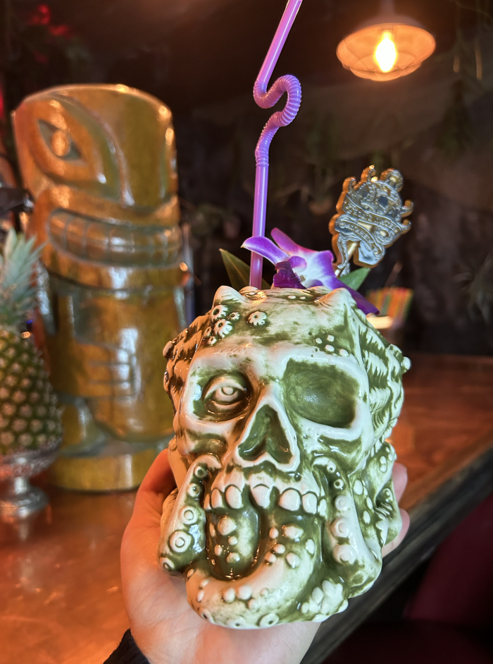 Image of a green cup shaped like a skull and octopus tentacle. The drink has a purple straw.